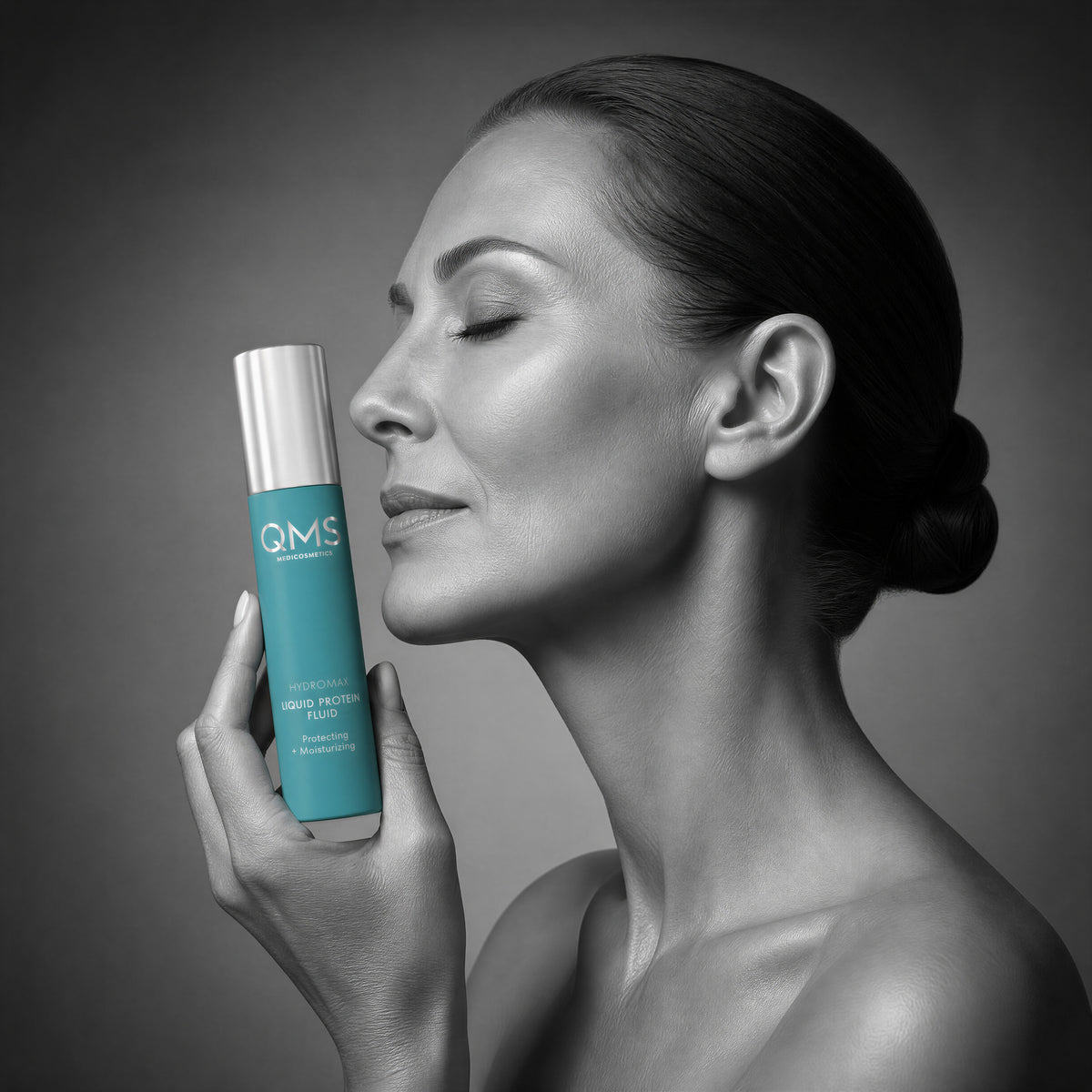 A woman in profile holds a turquoise cosmetic bottle with a silver cap near her face against a dark gray studio background. The bottle label shows the brand QMS.